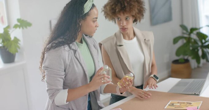 African American women with samples examining bottles at studio, using laptop for packaging layout