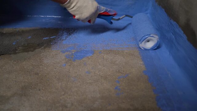 Close-up of a gloved hand painting blue waterproofing membrane on a concrete foundation corner.