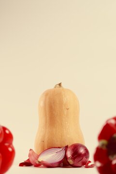 Vertical macro of butternut squash with red onion and halved onion with peeling husks on pastel yellow background. Blurry bell pepper in foreground. Organic vegetables with copy space on top for text.