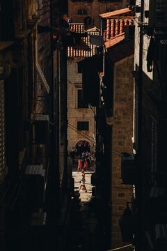 A dramatic vertical view of a narrow passageway descending toward the famous Stradun street in the Old Town of Dubrovnik, where sharp shadows frame sunlit tourists