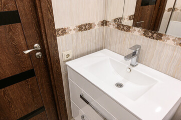 Fototapeta premium sleek white bathroom sink with a modern chrome faucet sits against beige tiled walls with decorative border, beside a dark wood door with metallic handle.