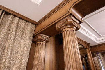 Rich wooden columns with fluted grooves and ornate capitals support a carved cornice  paired with patterned curtains and intricate ceiling moldings in a classic interior. © evgeniykleymenov