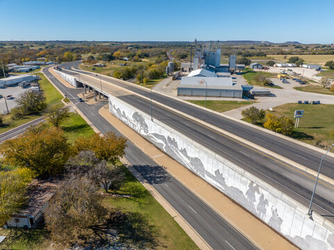 Aerial Drone View of Generic Gray Industrial Factory Building Surrounded By Trees Near Highway Road With Stone Wall Mosaic Mural in Comanche, Texas