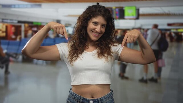 Young woman points both fingers to bare abdomen in busy airport terminal while smiling and wearing white crop top; travel confidence.
