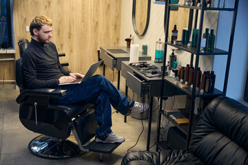 Male barber sitting in armchair and typing on laptop in hairdressing salon