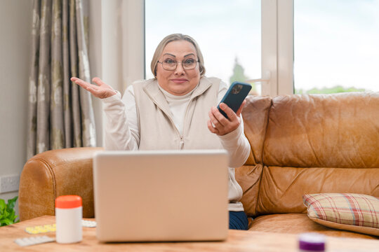 Woman sits on brown couch with laptop and phone while looking confused in a bright room