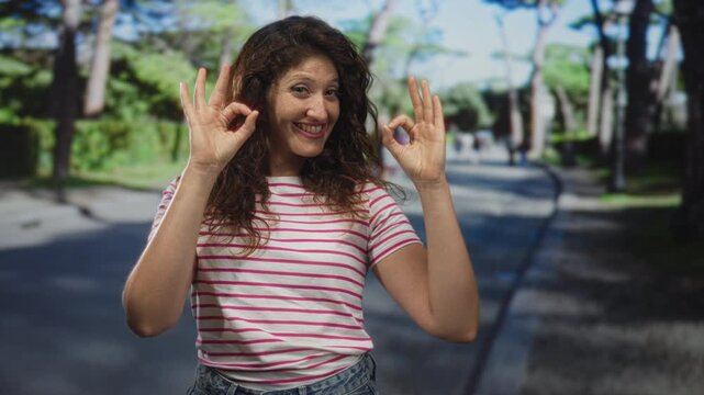 Woman shows ok sign with both hands on a tree lined street in a green park wearing pink striped t shirt and denim jeans; joyful optimism.