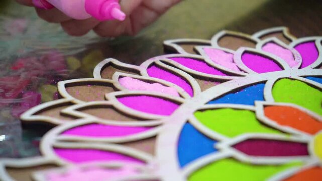 Slow motion macro close up shot of coloured powder being poured from a plastic bottle into a rangoli stencil template with vibrant floral pattern on the floor showing the traditional decoration of