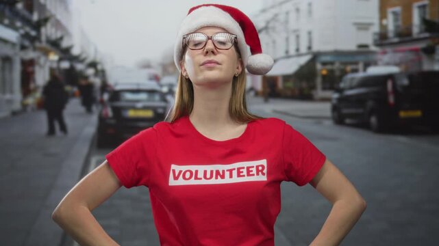 Woman wearing santa hat and volunteer tshirt on city street adjusting shirt with blurred cars and buildings visible; holiday generosity.