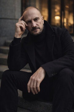 Portrait of a thoughtful bald man sitting on urban steps wearing a dark coat, moody evening city atmosphere with warm lights in the background.