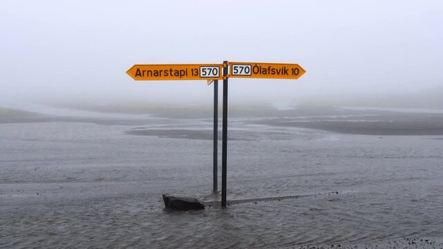 Icelandic road sign for Arnarstapi and Olafsvik submerged in flowing water during a foggy day