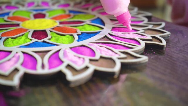 macro close up shot of coloured powder being poured from a plastic bottle into a rangoli stencil template with vibrant floral pattern on the floor showing the traditional decoration of hindu festivals