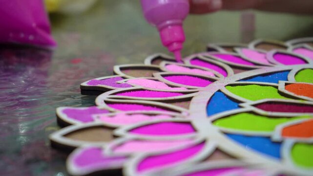 Moving forward shot showing colored powder being poured using plastic bottle with nozzle into stencil template with vibrant floral pattern on the ground showing hindu indian festival of holi diwali