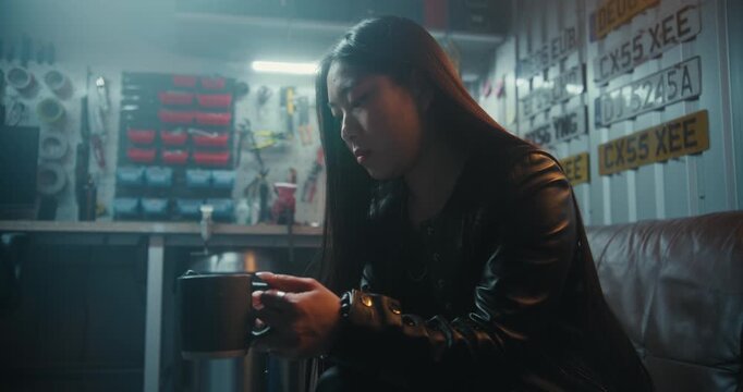 Young Asian Woman With Long Hair in Leather Jacket Drinks Hot Coffee While Scrolling Social Media on Smartphone. Concept of Motorcycle Lifestyle and Digital Communication in Professional Workshop.