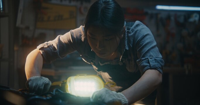 Dressed in Work Overalls and Protective Gloves, Young Female Leans Over Open Hood, Using Handheld Lamp to Illuminate Hard to Reach Areas of Engine. Expert Observation and Identifying Potential Faults.