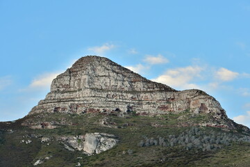 The rocky peak of Lion's Head mountain stands against a clear blue sky with scattered clouds in Cape Town, South Africa. The sandstone cliffs displays the natural vegetation of the iconic landmark.