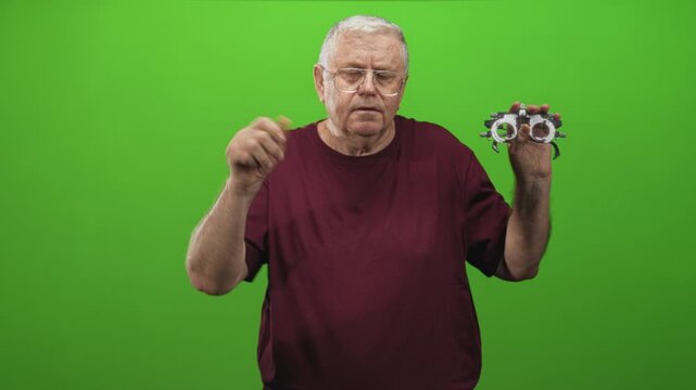 Elderly man holds trial frame with raised hand during eyeglass examination against green screen in studio; concern.