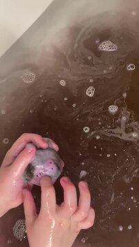 Children hands lower a bath bomb into water. The bath bomb begins to fizz, foam, and dissolve, coloring the water. The scene takes place indoors during a bathing routine.
