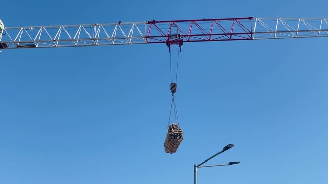 Tower crane carrying bundled load across skyline, suspended materials swing past streetlight, clear blue sky backdrop, transit