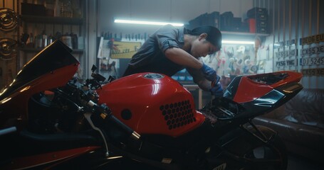 Naklejka premium Professional Female Rider Detaching Battery Cables Using Screwdriver In Workshop. Asian Woman Mechanic in Overalls Works on Electrical Components of Red Motorbike. Concept of Smart Garage Lifestyle.