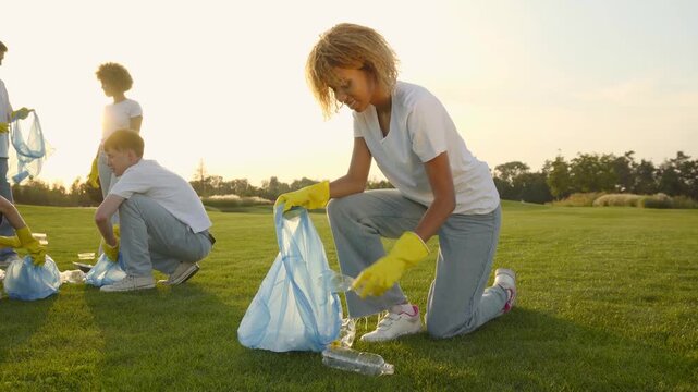 Multiracial group of young volunteers at smiling woman with Afro Hair wearing yellow gloves and filling blue trash bags while cleaning a sunny park, conveying teamwork, community service 4k