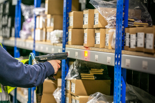 Warehouse operative using a handheld barcode scanner to scan a location label on a picking shelf, with stocked inventory visible on surrounding shelves