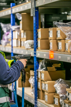 Warehouse operative using a handheld barcode scanner to scan a location label on a picking shelf, with stocked inventory visible on surrounding shelves
