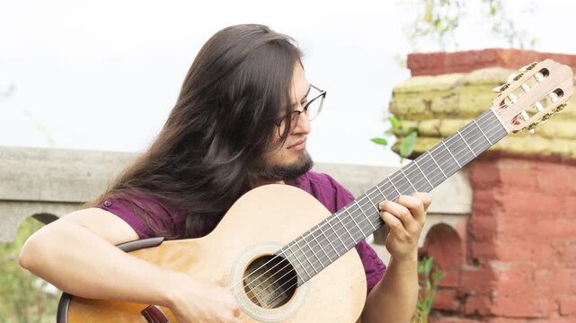 Horizontal medium close-up of a male musician with long dark hair, a goatee, and glasses. He is wearing a purple shirt and is captured with a serene, focused expression while playing a classical acous