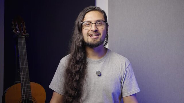 Medium shot of a man with long, wavy dark hair, a goatee, and glasses. He is wearing a simple grey t-shirt with a small lapel microphone attached.
