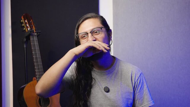 Horizontal medium shot of a man with long, wavy dark hair, glasses, and a goatee, smiling warmly at the camera. He is wearing a grey t-shirt with a small lapel microphone attached.