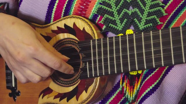Horizontal close-up shot focusing on a musician's hands playing a charango, a small Andean stringed instrument. The performer is dressed in a traditional poncho with vibrant, multi-colored geometric p