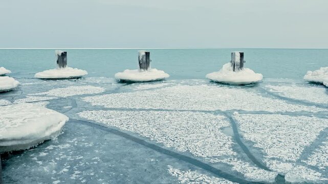 Wooden posts standing in icy water, surrounded by patches of snow and ice on the surface. The view captures the quietness of a winter day by the water.