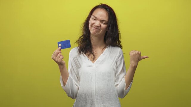 Woman holding creditcard in one hand with thumb pointing to self and smirk, wearing white blouse in studio; skepticism doubt money.