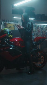 Stylish Asian Woman in Leather Gear Using Phone Map While Sitting on Red Sport bike in Garage. Young Female Focused Scrolling and Enlarging Screen See Road Details in Private Workshop. Vertical Shot.