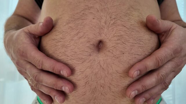 Close-up of an overweight man holding his belly from below and slapping stomach fat with both hands. Concept of weight gain, obesity and unhealthy lifestyle.