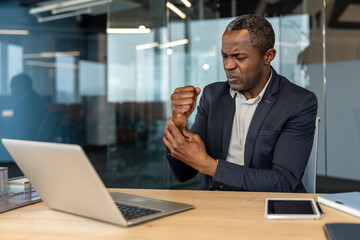 Obraz premium Mature man experiencing discomfort and pain in his wrist while working at his desk with a laptop, indicating potential musculoskeletal problems or carpal tunnel syndrome from prolonged computer use