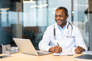 Doctor in lab coat and stethoscope smiling at desk, writing on a clipboard with laptop and tablet...