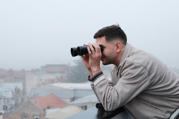 Naklejka premium Young man leaning on balcony railing and looking through binoculars over foggy city. Concept of observation, urban exploration and discovering the city view.