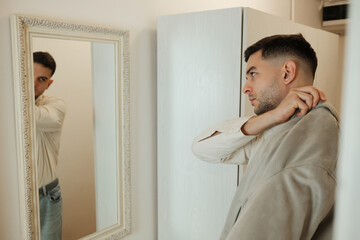 Fototapeta premium Young man looking at his reflection in mirror while adjusting clothes before leaving home. Morning routine and daily lifestyle concept.