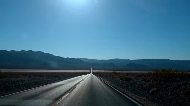 Perspective view of long straight desert highway under bright sun. Symmetrical shot of empty asphalt road leading to distant mountains in an arid desert environment under blue sky.
