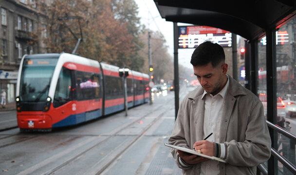 Young man writes in a notebook while waiting at a tram stop in the city. Public transport and productivity.