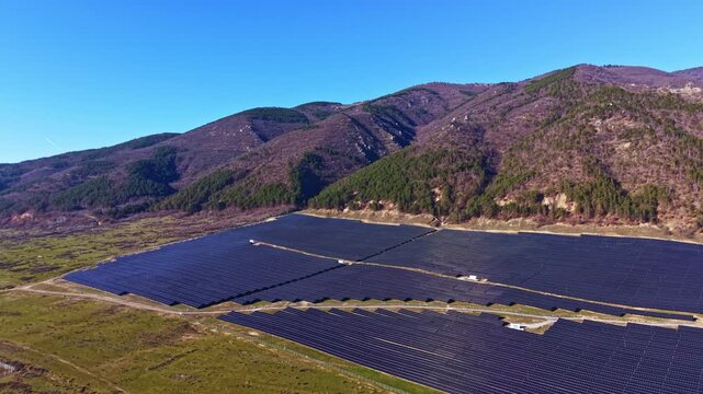 Solar panels spread across a wide area on flat land, surrounded by hills under a clear blue sky. The scene shows the use of renewable energy in a rural setting.