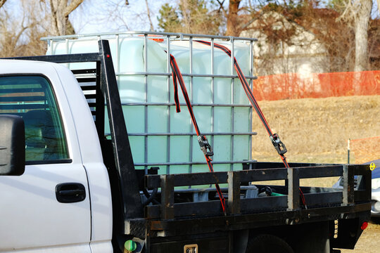 Large plastic IBC water tank secured with ratchet straps on a pickup truck. Utility vehicle transporting liquid container, industrial water storage cube for construction, farming or landscaping