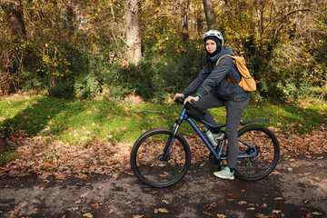 Fototapeta premium Woman riding bicycle along autumn park path surrounded by golden trees. Active lifestyle and eco friendly transport concept.