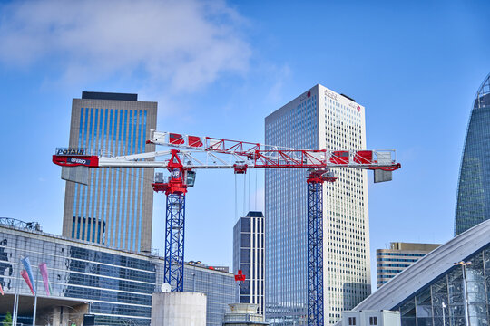 A red and white Potain construction crane stands prominently in the foreground against the modern skyline of La D&eacute;fense business district in Paris, France. The crane features visible branding