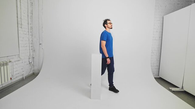 Confident Caucasian guy in T-shirt and trousers is walking around a pillar in a studio filmed in slow motion. Young adult bearded man in glasses is going on a circle way. Pattern of human movement