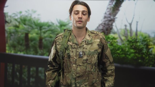 Soldier man in camouflage uniform wearing dogtag with hand to chest on building balcony, backpack strap visible; duty homecoming.