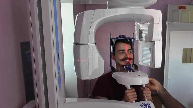 Patient in panoramic xray scanner smiling broadly, seated upright with chin support and hand grips, operator initiating imaging sequence in contemporary dental