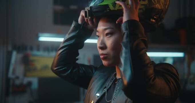 Close Up of Young Asian Woman Puts on Black Helmet With Green RIDE Graphics, Fastens Chin Strap, Lowers Visor For Eye Protection. Female Rider Putting Helmet in Garage. Motorcycle Safety, Preparation.