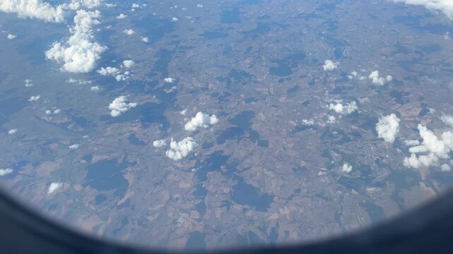 Aerial view from an airplane window looking down at patchwork agricultural fields and scattered clouds over rural Europe.